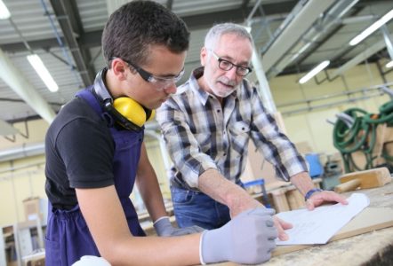 An apprentice and a teacher discussing plans in a workshop