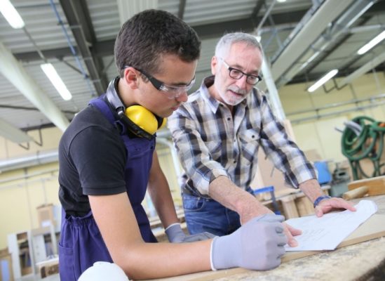 An apprentice and a teacher discussing plans in a workshop