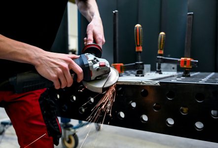 Person using an angle grinder to cut metal on a workbench, with sparks flying and tools clamped in place in an industrial workshop