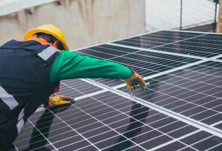 Worker wearing a hard hat and gloves installing solar panels on a rooftop, tightening a mounting fixture on the panel frame