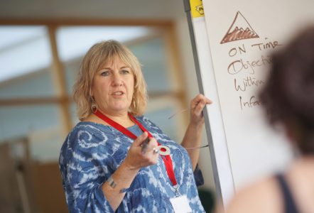 A teacher or trainer wearing a red lanyard gestures while explaining something on a flip chart during a classroom or workshop session.