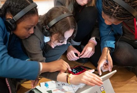**Alt text:** Four secondary school students wearing headphones huddle around a tablet and audio equipment, working together on a hands-on digital activity. Papers and learning materials are spread across the floor as they concentrate on the task in a museum learning space. **Shorter version:** Four students wearing headphones collaborate around a tablet during a STEM learning activity in a museum setting.