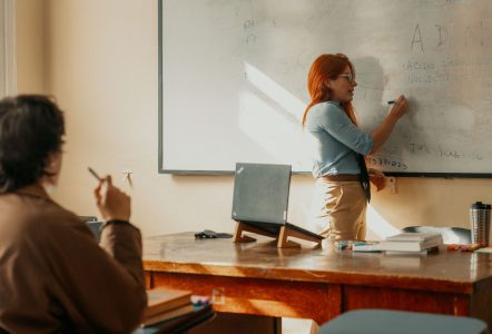 A woman writes on a whiteboard while presenting to a seated participant in a sunlit classroom or workshop setting.