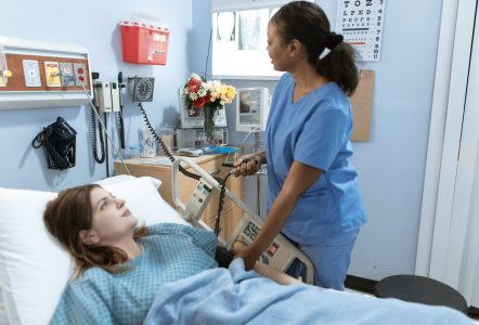 A healthcare worker in blue scrubs checks on a patient lying in a hospital bed in a clinical ward.