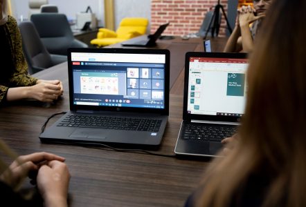 People gathered around laptops during a hybrid meeting, with a video call and presentation displayed on screen.