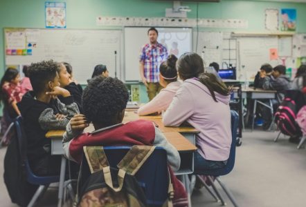 Teacher addressing pupils seated around tables in a classroom.