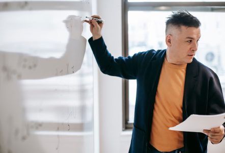 A teacher holding papers and writing on a whiteboard in a bright classroom.